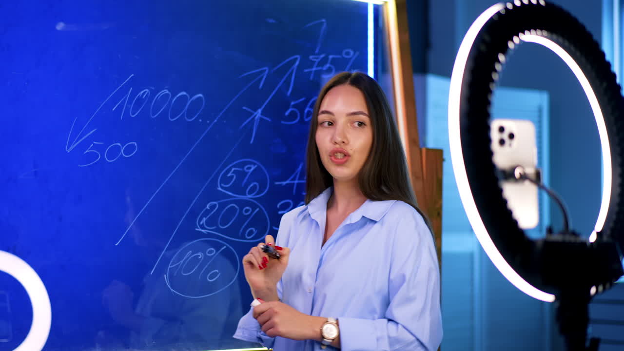 Young woman teaching finance online. A young woman writes on a glass board while explaining financial concepts during an online class in a bright studio