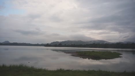 Conduciendo-Por-Un-Tranquilo-Lago-Brumoso-Con-Montañas-Al-Fondo