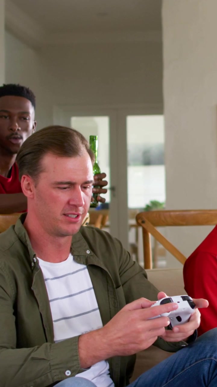 Vertical video: Holding green-bottled helmet, pals teasing player on couch at home with gamepad