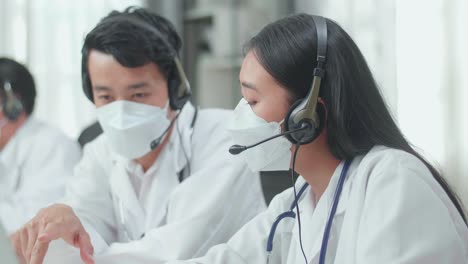 Close Up Of An Asian Man And A Woman Doctors In Headsets And Masks Working As Call Center Agent Discussing Work While Their Colleague Is Speaking With Customer On The Call At The Office