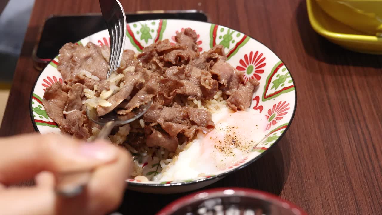 A person uses a spoon and fork to eat a bowl of sliced beef, rice, and a boiled egg in a brightly lit restaurant setting