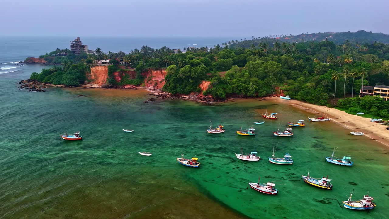 Weligama köyü plajı açıklarında, sığ turkuaz suda duran balıkçı tekneleri, tropikal sahil boyunca uzanan kırmızı kayalıklar, palmiye ağaçları ve hafif dalgalarla.