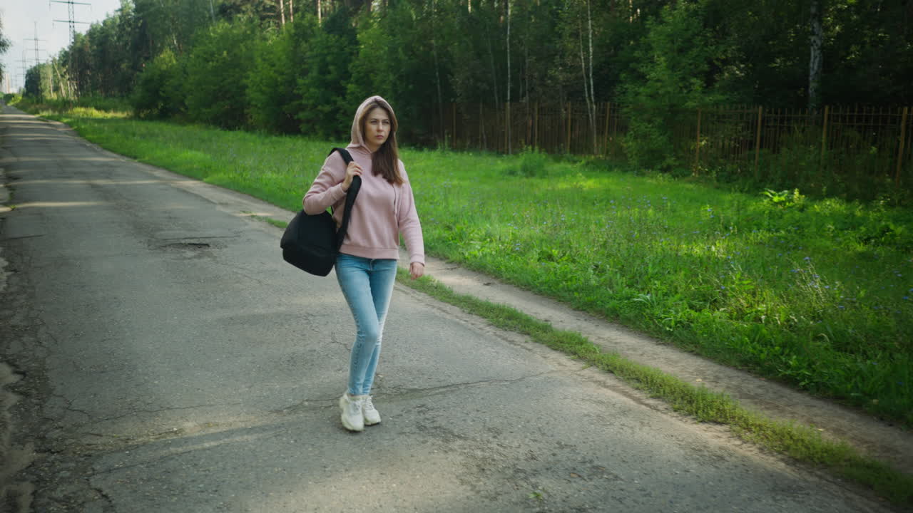 Beautiful woman in pink hoodie walking along quiet paved forest road, holding black bag strap with one hand, wearing jeans and sneakers, surrounded by lush green grass and tall trees under clear sky