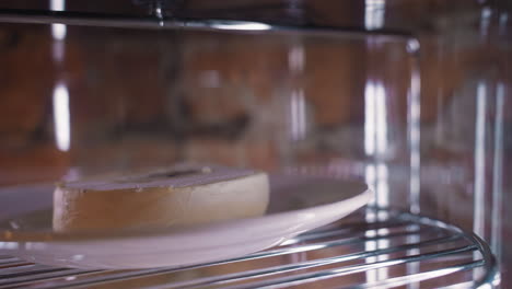 creamy dessert cake with smooth frosting placed on white plate, rotating on metal shelf inside transparent display fridge under soft lighting, highlighting texture and chilled presentation