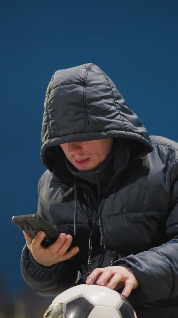 un hombre sentado en un banco con una pelota de fútbol entre las piernas, devuelve su teléfono a su bolsillo, y comienza a girar la pelota de una manera lúdica