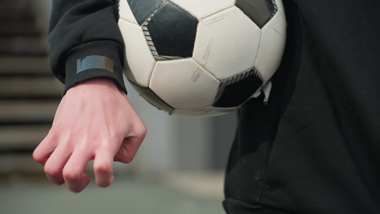 Close-up of an individual holding a worn soccer ball under his arm, as he clasped hand, with a blurred view of a staircase behind