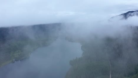 aerial passing through fog and clouds high over scenic lake in forest