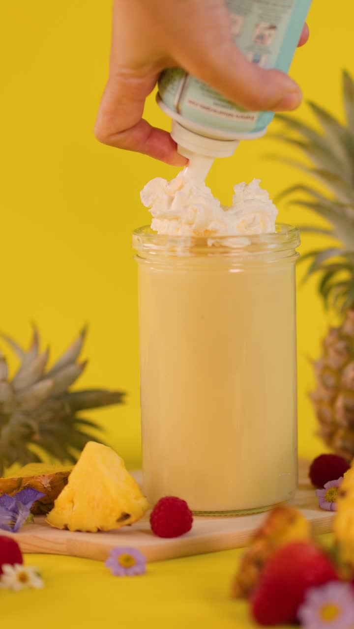 Una mano guarnisce un frullato all'ananas con panna montata e frutta su uno sfondo giallo vivace, scatto statico.