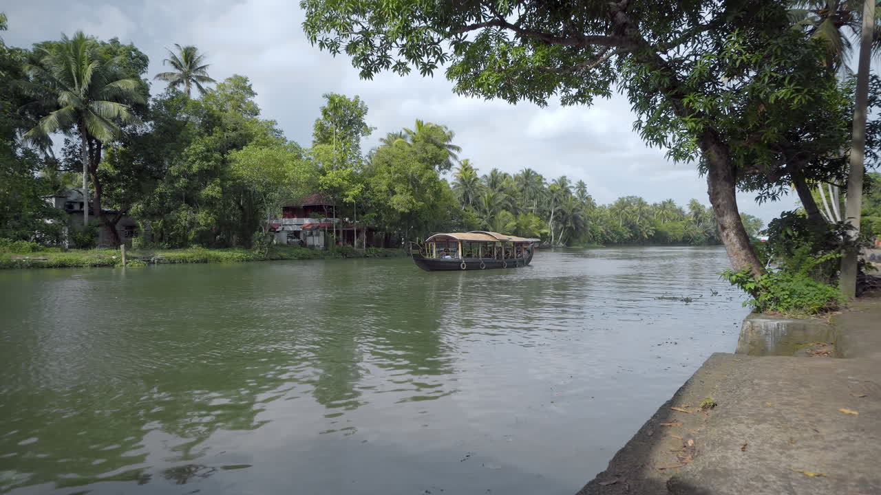 un barco shikara o kettuvallam pasa por el pueblo en los remansos de kumarakom, kerala, sur de la india en un día soleado con un exuberante fondo verde de cocoteros y un cielo azul nublado