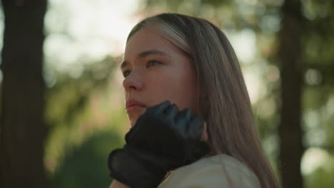 beautiful young woman resting chin on hand, gazing thoughtfully into distance with sunlight creating warm halo effect around her face, blurred greenery in background