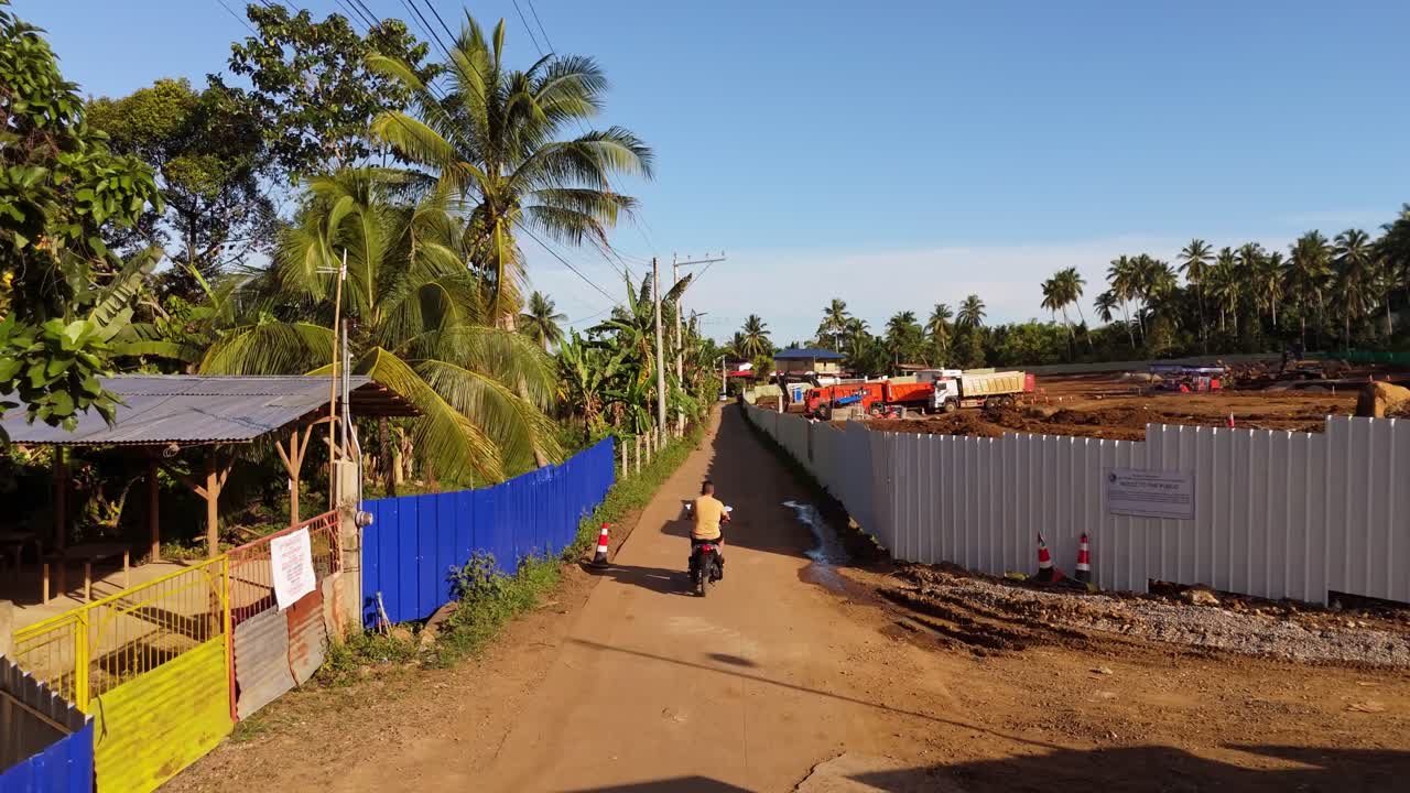 A man rides his motorbike along a sunlit village road in the Philippines, flanked by palm trees and local fencing near a construction site — showcasing daily life in a tropical island community