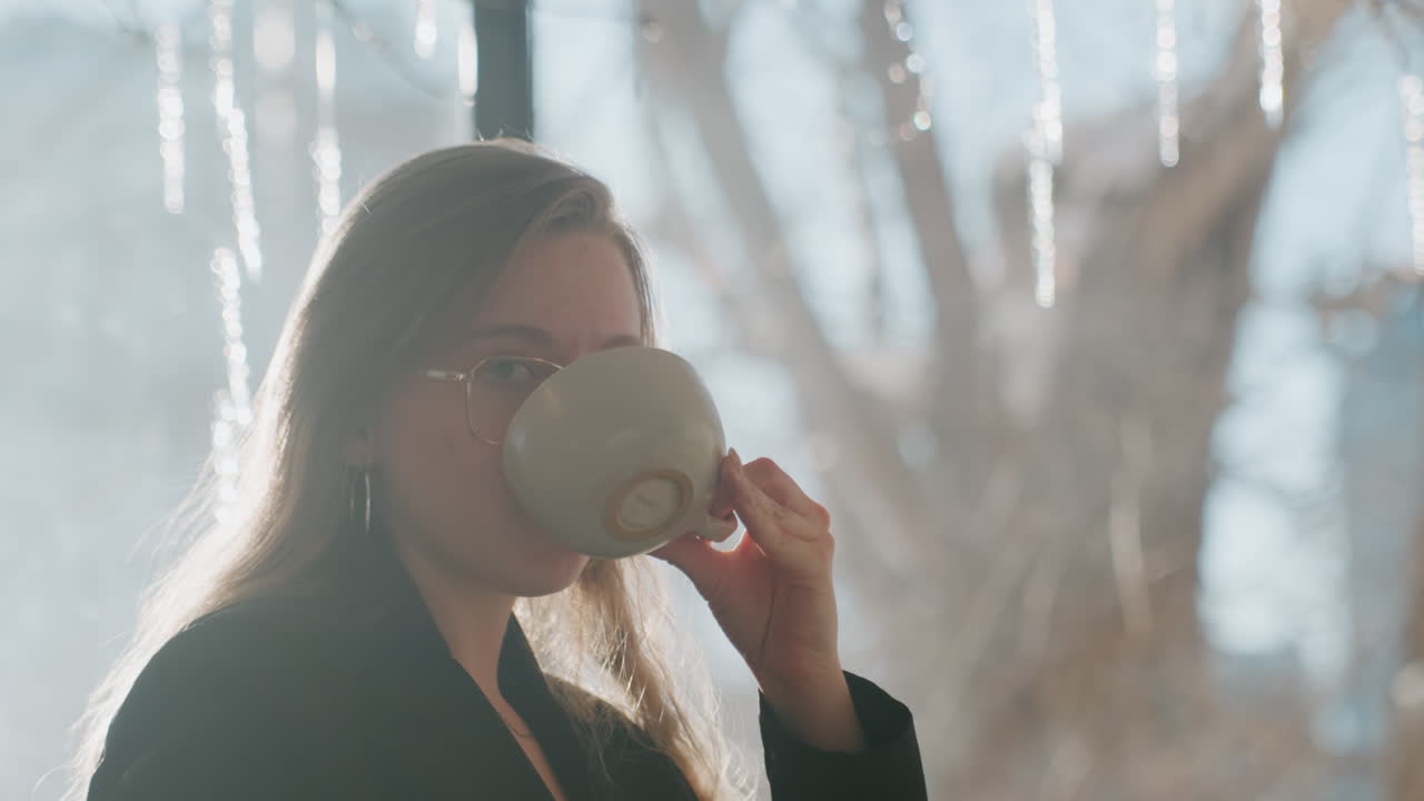 Hvid kvinde i eftertænksomt portræt ved vindue med istapper, holder kop og kigger eftertænksomt, kantlys fremhæver hår og briller, fredfyldt kreativ atmosfære, blød vinterbaggrund.