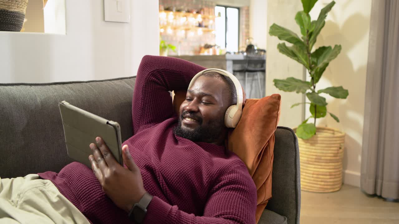 African American man reclining on gray sofa at home swiping tablet and smiling with headphones