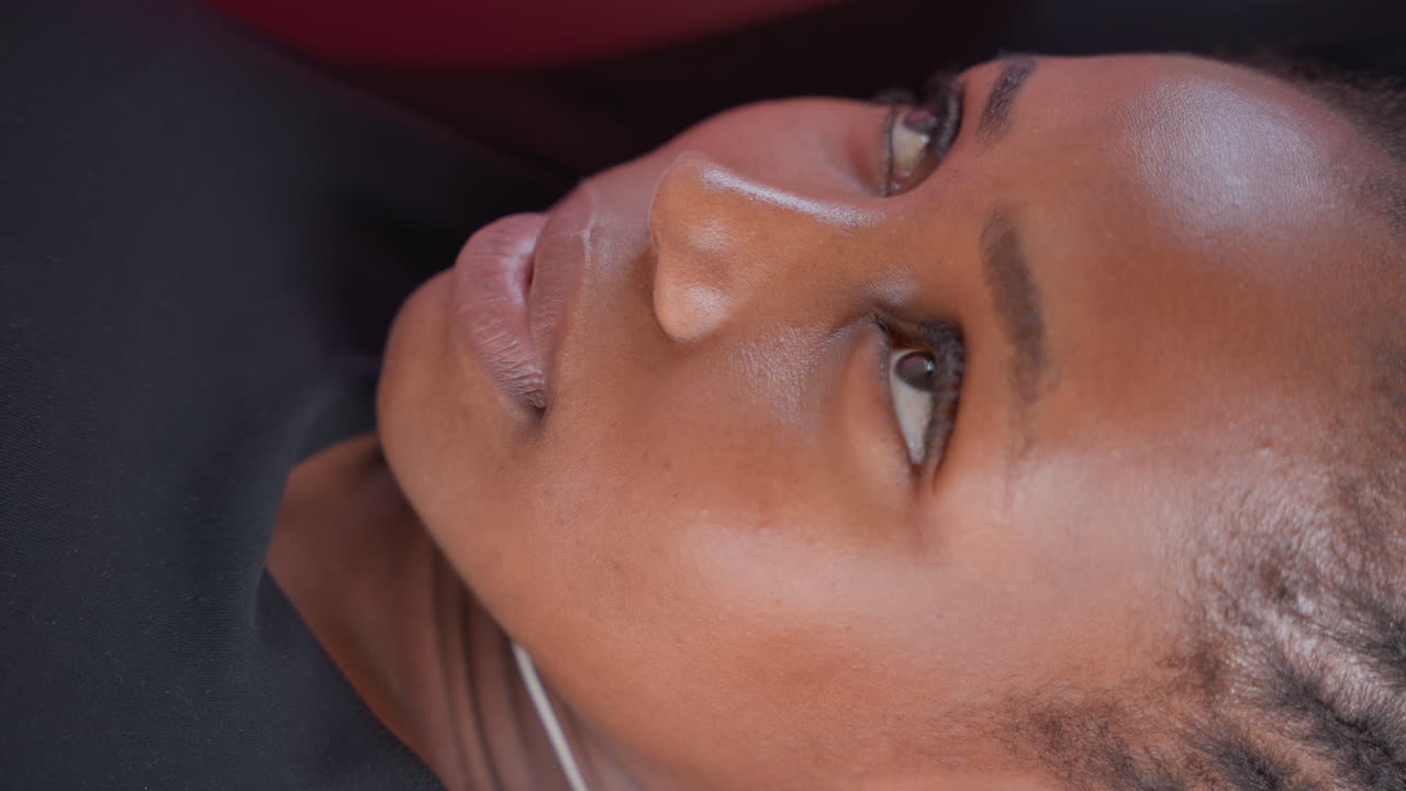 Close-up de mulher negra deitada calmamente, pele brilhante, tranças nagô, fone de ouvido branco, iluminação suave de estúdio, expressão pensativa, cliente de spa descansando durante momento de cuidados com a pele, detalhe íntimo da textura, bem-estar.