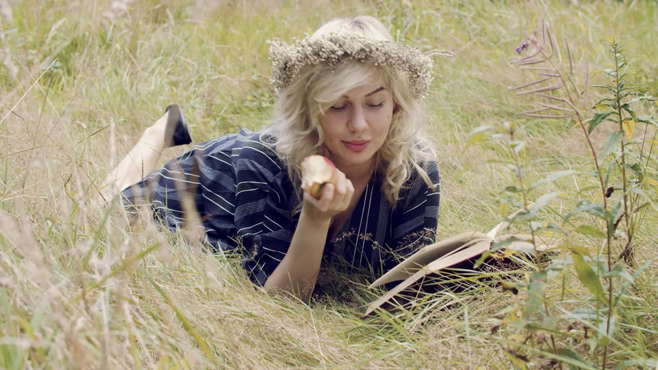 Attraktiv blond kvinde ligger på en græsmark, spiser et æble og læser en bog
