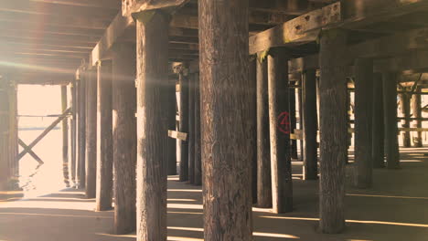 Atmospheric-beautiful-scenery-underneath-a-dock-in-Charleston-Oregon-with-sun-flare,-rays-and-shadows-at-sunrise,-boats-in-the-background