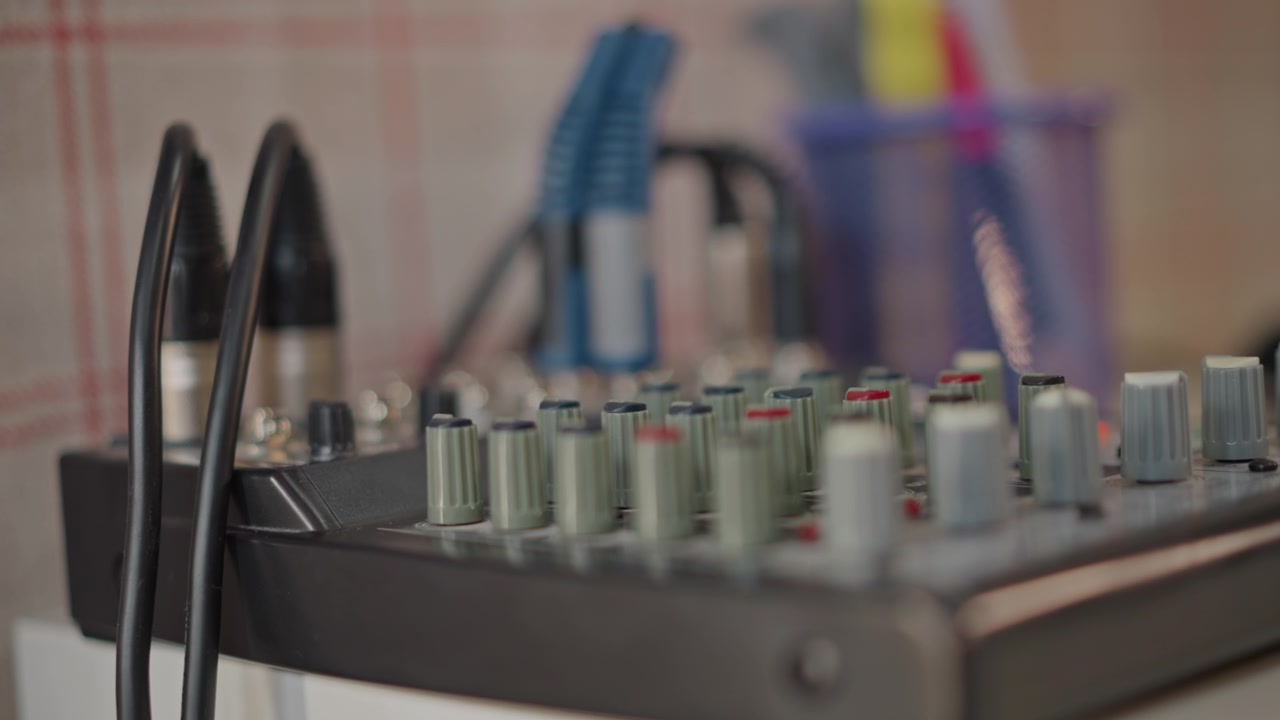 Mixerpult med tilsluttede kabler og jackstik, studerende musiker forbinder kanaler, mens instruktør forbereder niveauer til skoleprøve, neutral ternet baggrund og lav dybdeskarphed fremhæver udstyr.