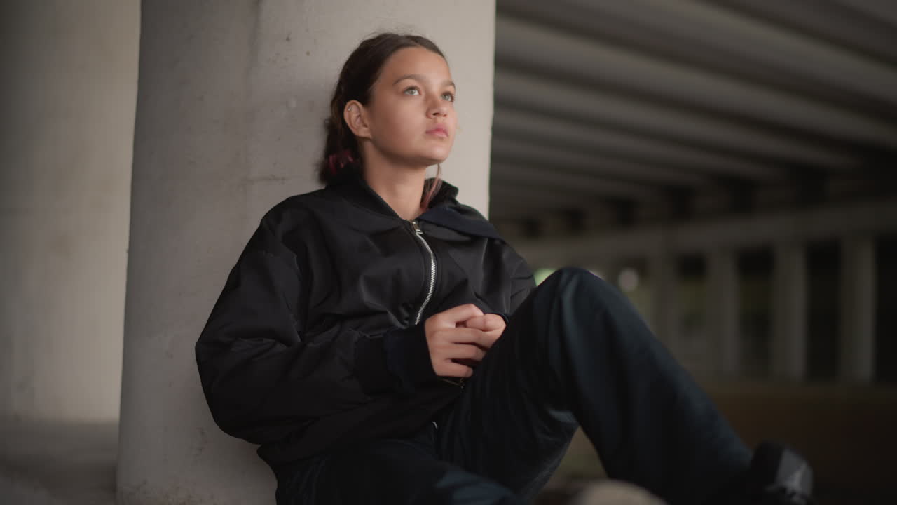 Jovem mulher encostada em pilar de concreto, skatista descansando com equipamento aos pés e telefone na mão, passagem subterrânea escurecida pela chuva, luz cinematográfica suave e humor introspectivo capturando um momento de descanso urbano.