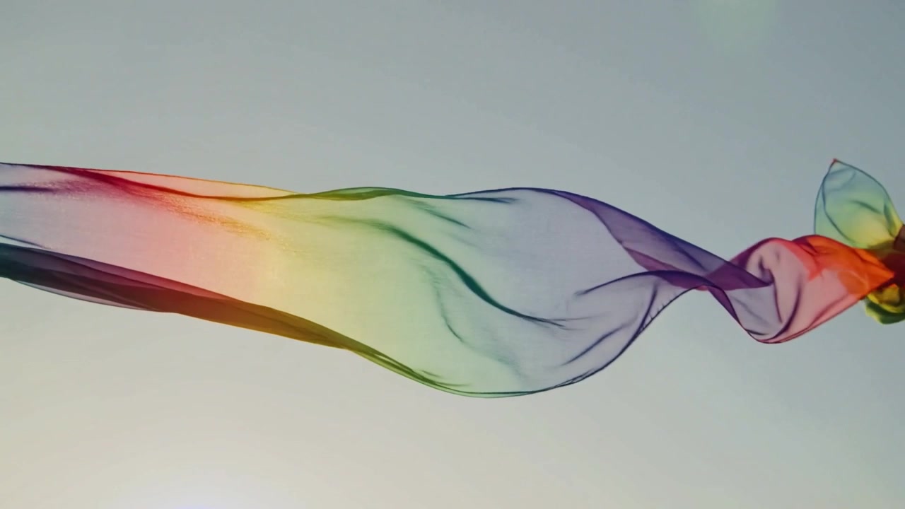 Drifting long translucent rainbow ribbon across clear sky, breeze causing unfurling and sun glare