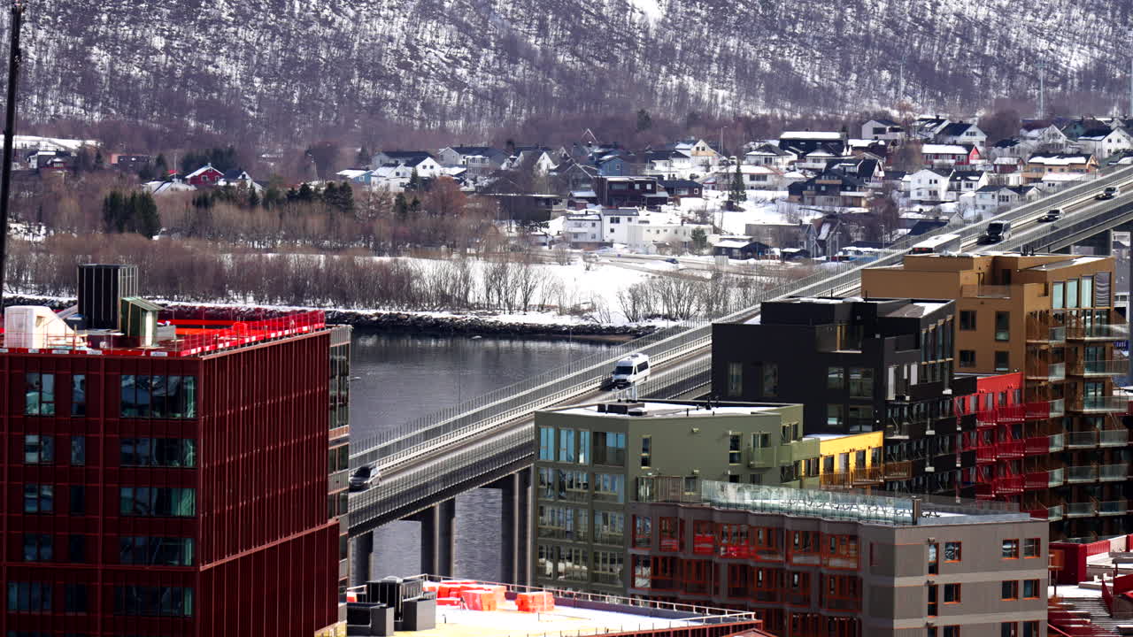 Tromsø Bridge stretching across the fjord, with modern colorful buildings, vehicle crossings and snowy residential hillside in the background during early spring daylight