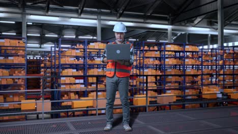 cuerpo lleno de ingeniero masculino asiático con casco de seguridad trabajando en una computadora portátil mientras estaba de pie en el almacén con estantes llenos de bienes de entrega