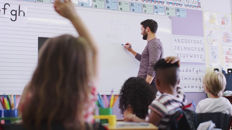 Male Teacher Standing At Whiteboard Asking Elementary Pupils Question In School Classroom
