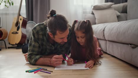 a little brunette girl in a pink dress lies on the floor and draws with her father, a brunette man in a checkered shirt, on a sheet of paper using multi-colored felt-tip pens in a modern apartment
