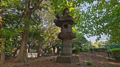 a weathered traditional japanese stone lantern in tokyo's ueno park. sunlight filters through lush trees, highlighting fallen leaves and creating a tranquil, historic atmosphere.