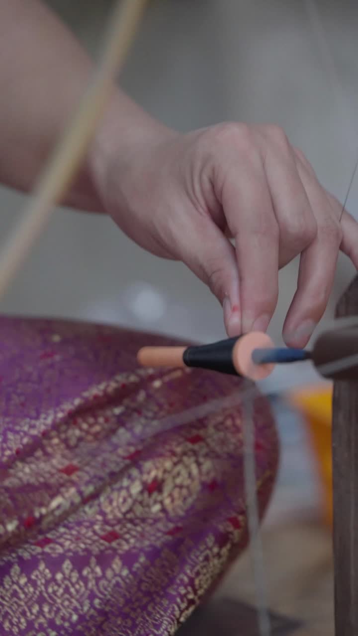 Close up slow motion silk thread wrapping around a bobbin with traditional patterned fabric in background artisan weaving process silk production workshop in Thailand vertical format