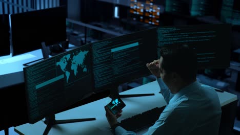 side view of happy asian man developer looking at code on mobile phone while write code with multiple computer screens in the office