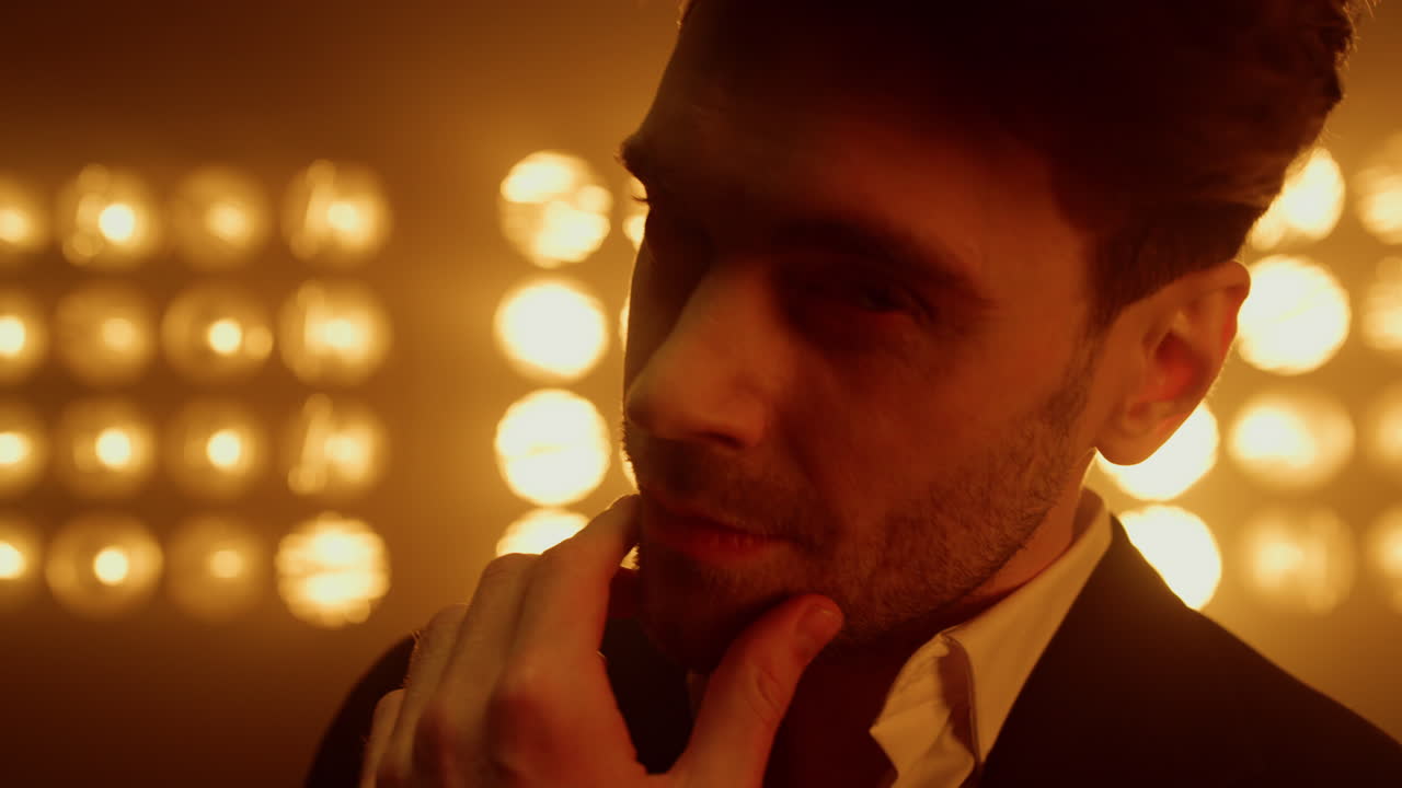 섹시한 남자가 얼굴을 만지며 무대 위에서 카메라를 쳐다보며 가까이 다가옵니다. 장면에서 포즈를 취하는 남자.