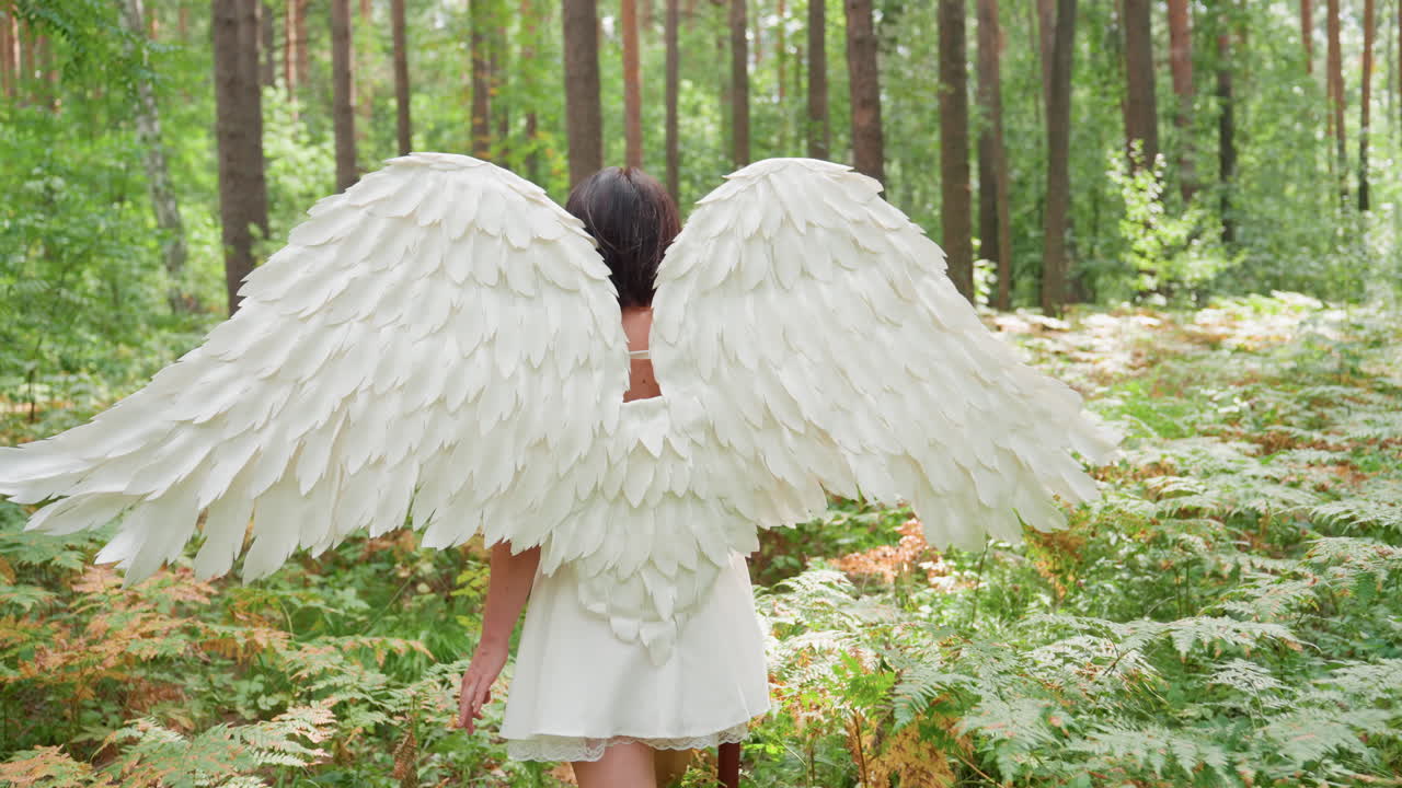 Back view of angel lady in white dress with large feather wings walking slowly through forest holding wooden staff surrounded by sunlight filtering through green trees