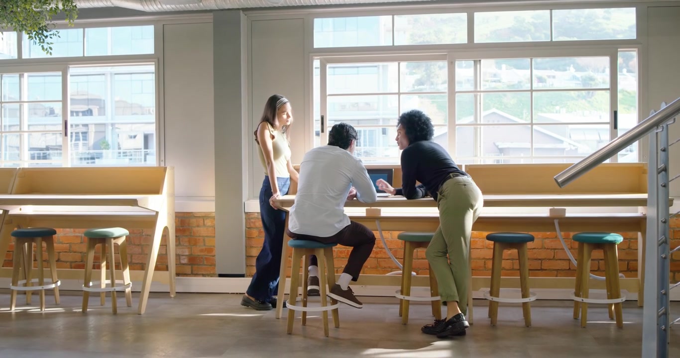Diverse team watching woman walking across open office carrying laptop and sharing work on counter