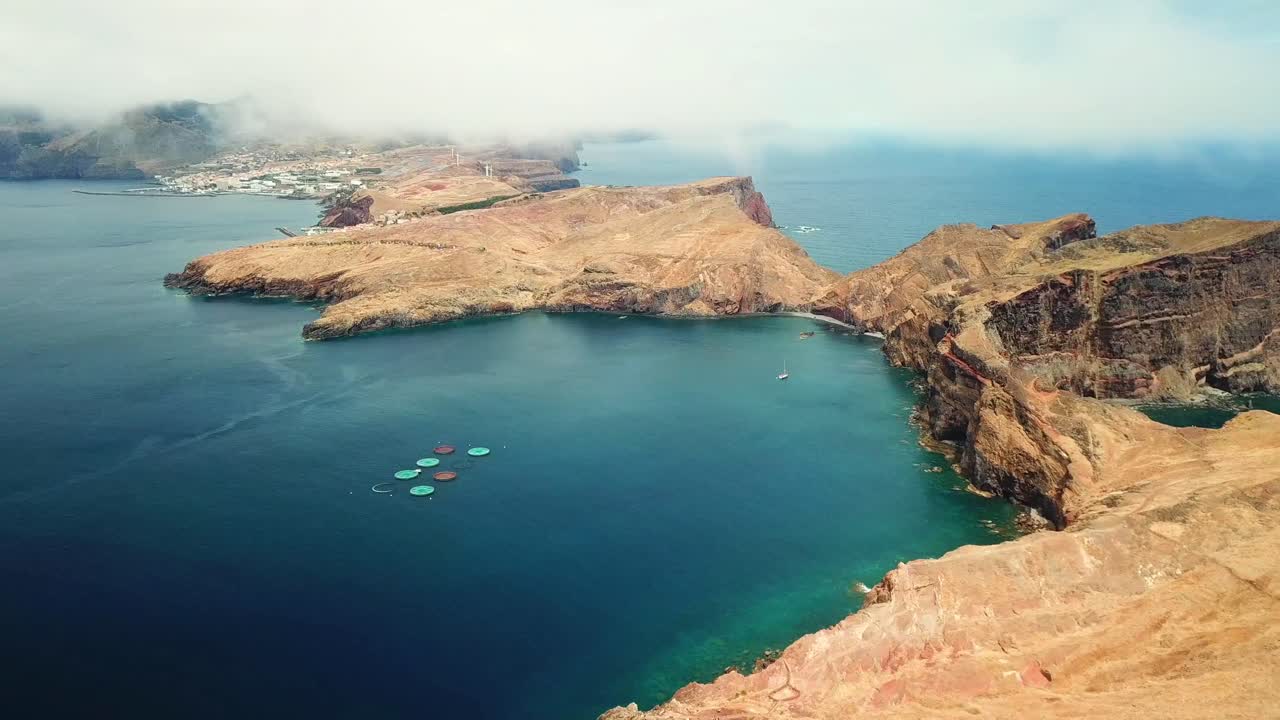 Aerial view of Ponta de São Lourenço, Madeira Island, showcasing fish farms in the foreground, with the rugged coastline, blue waters, and a distant town under a cloudy sky drone establishing shot