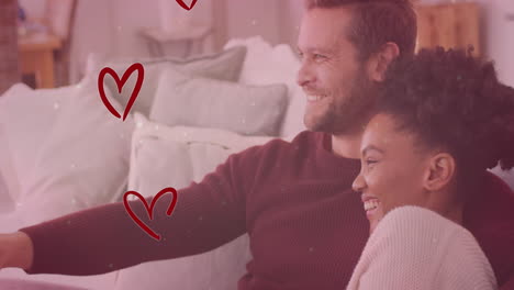 animación de corazones y puntos de luz sobre una pareja diversa abrazándose en el sofá y viendo la televisión