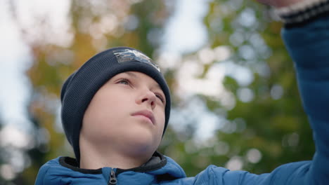 close-up of boy in black beanie and blue jacket thoughtfully observing dry leaf before dropping it, standing amidst vibrant greenery, blurred background with colorful autumn foliage