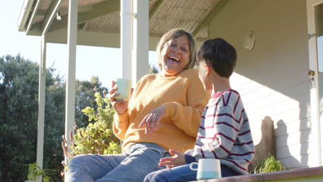 Feliz-Abuela-Birracial-Y-Nieto-Sosteniendo-Tazas-Sentados-En-La-Terraza,-Cámara-Lenta