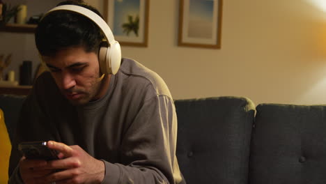 young man spending evening at home sitting on sofa wearing wireless headphones streaming music from mobile phone 1