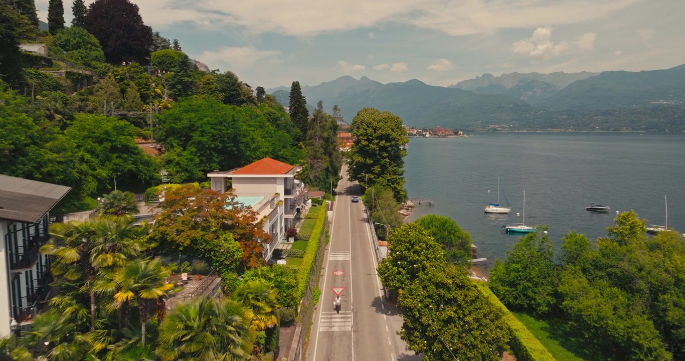 Drone vola sopra la strada lungolago del Lago Maggiore, rivelando ville italiane storiche, barche che riposano lungo la riva, traffico di passaggio e spettacolari montagne alpine