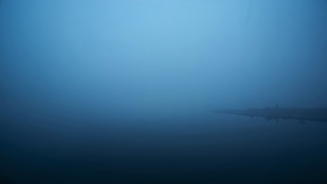 Una telecamera che riprende un lago avvolto dalla nebbia con acqua calma, nebbia densa e una riva sfocata sulla destra.