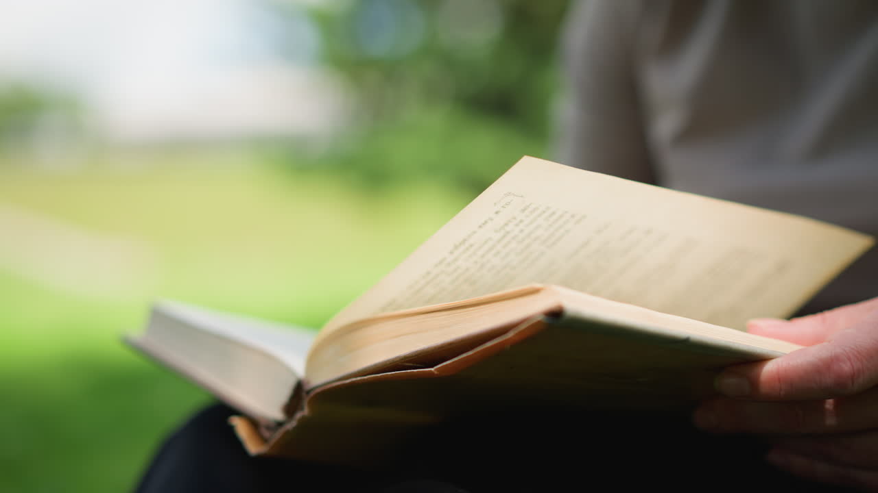 Close up partial view of lady flipping through old book pages with hand gesture in soft focus, green blurred background of park creating peaceful atmosphere of reading, curiosity