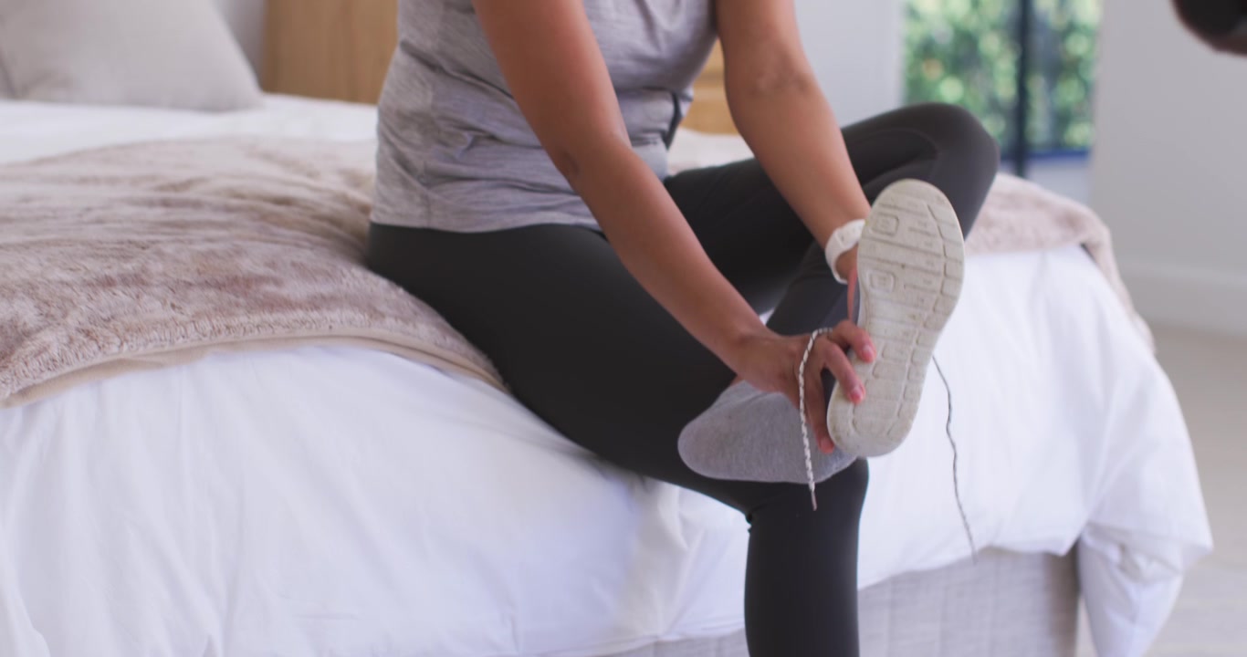 Donna afroamericana seduta sul letto che infila il piede in una sneaker grigia allacciando i lacci, preparandosi per l'allenamento