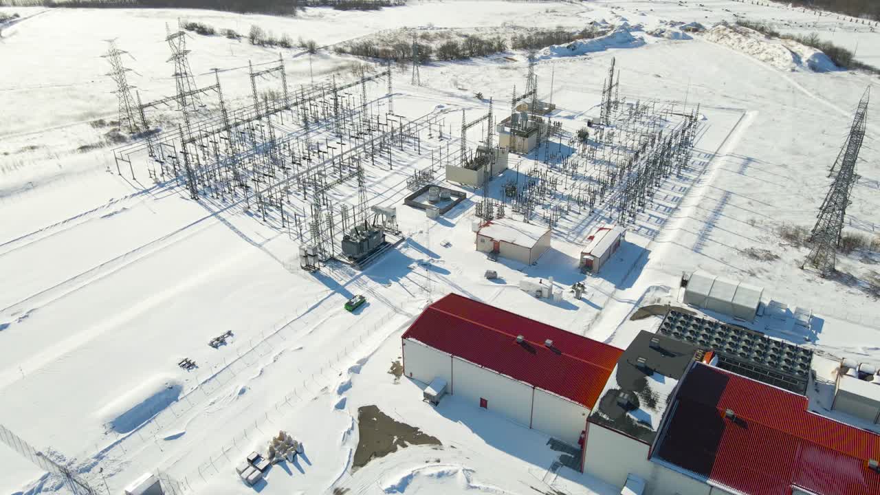 Dron letící pozpátku nad elektrickou rozvodnou připojenou k modernímu datovému centru s bateriovým úložištěm a chladicími systémy v zasněžené zimní krajině