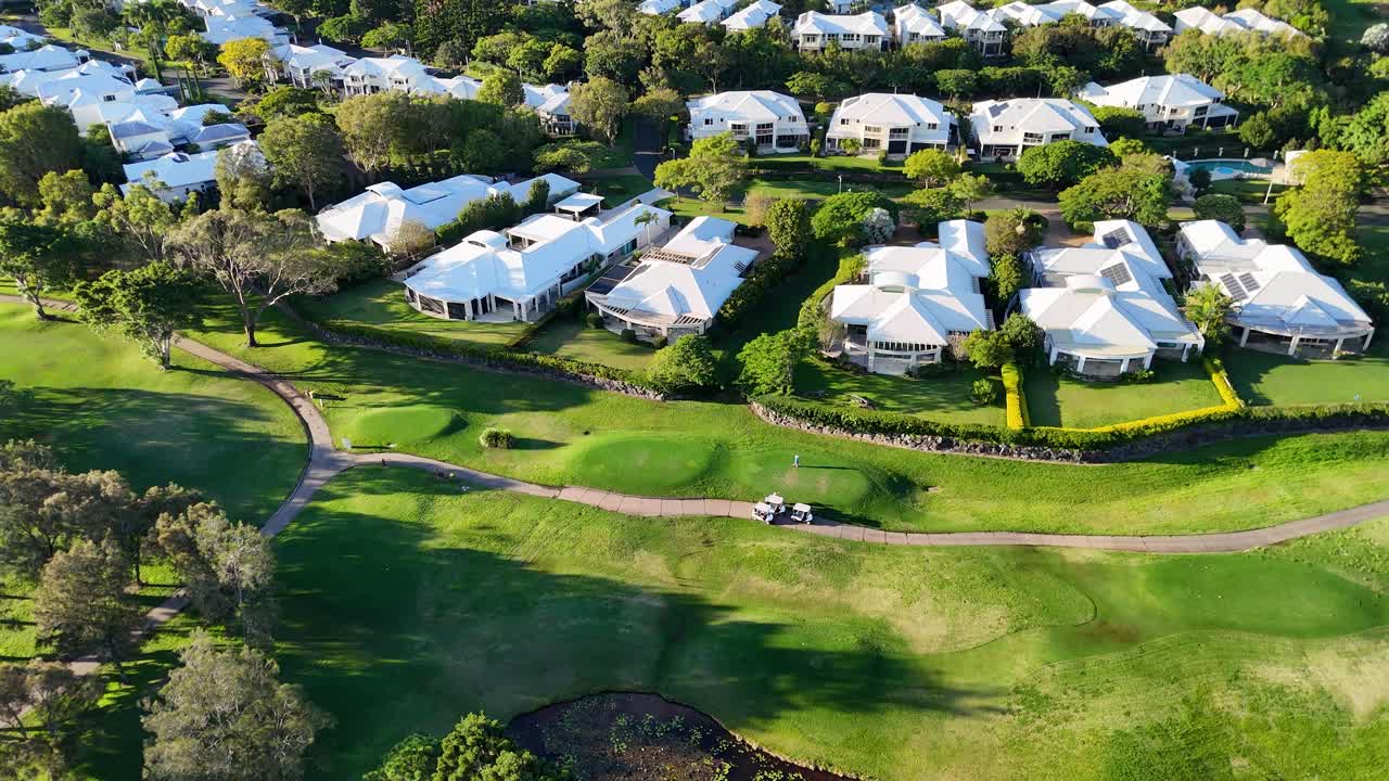 Drone footage captures a serene golf course with houses, trees, and a lake during golden hour