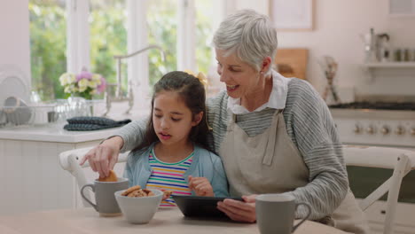 grandmother and little girl using tablet computer watching online entertainment eating cookies in kitchen happy granddaughter enjoying sharing weekend with granny browsing internet together