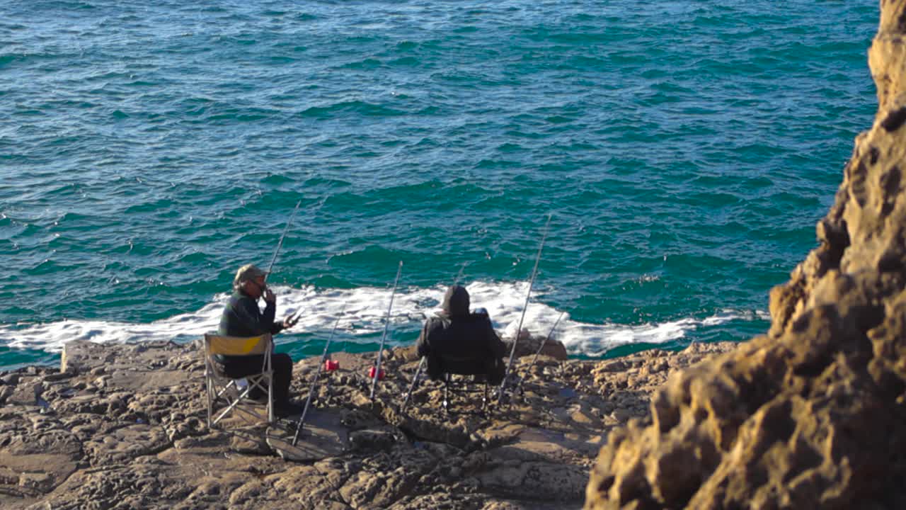 Two elderly fishermen sit high above the ocean at Boca do Inferno in Portugal while fishing and talking together as deep blue water moves below on a bright sunny day