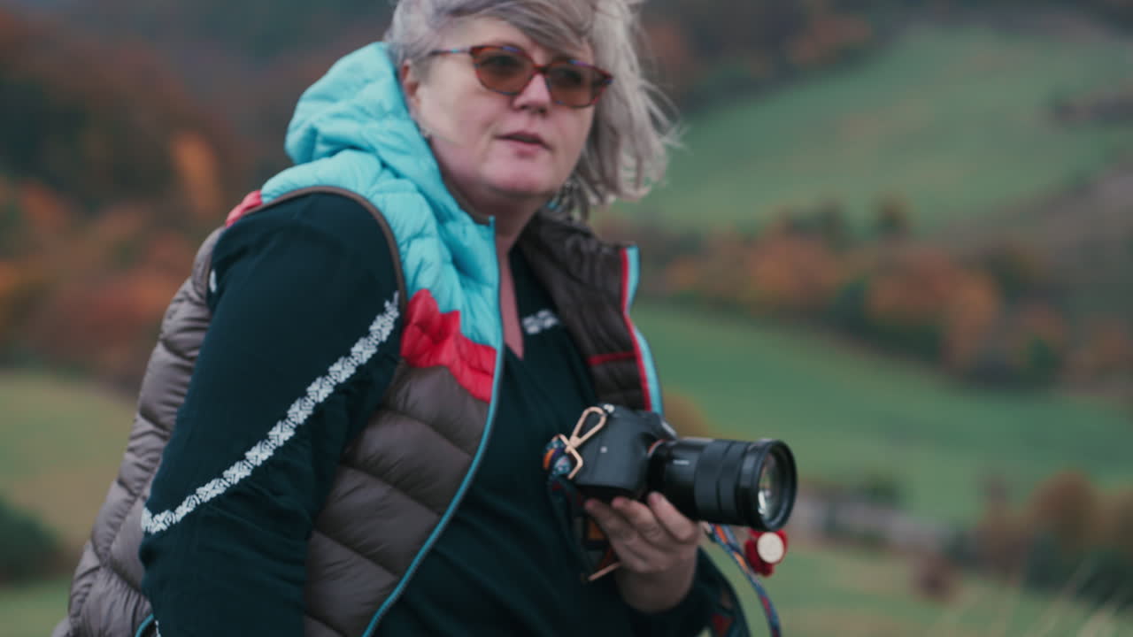 Una donna guarda la sua fotocamera e stabilisce un contatto visivo diretto durante un freddo e ventoso autunno, immersa in una natura colorata con alberi dalle foglie arancioni, di giorno.