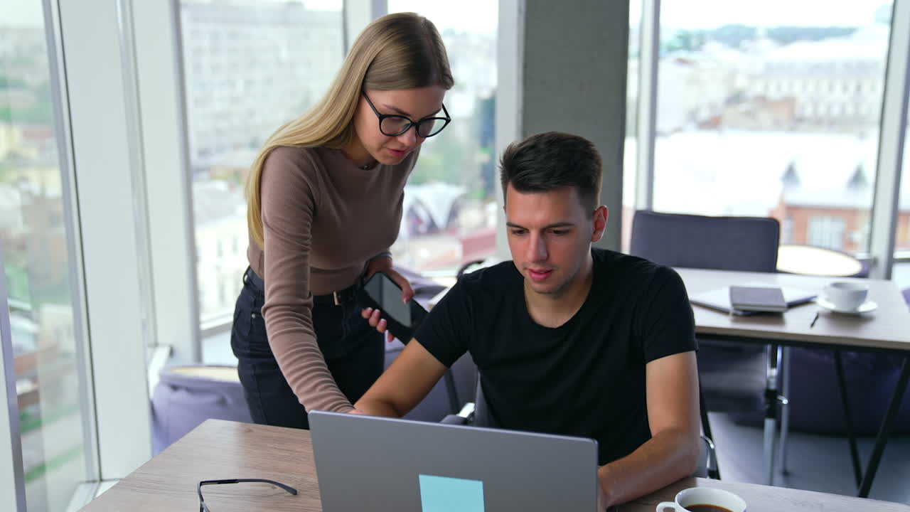 Lady wearing glasses stands near man working at laptop. Woman points at the screen commenting it. Panoramic windows in blur at backdrop.