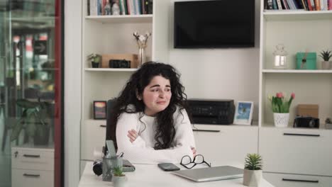 the young brunette woman with long curly hair sits at the desk in the office; she is very sad and worried about her work