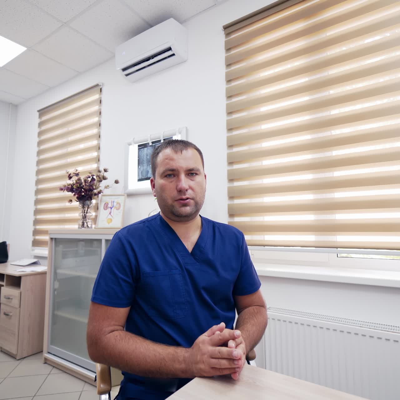 Online consultation. Professional male doctor in blue uniform sitting at table looking at camera and talking about health treatment in medical office.